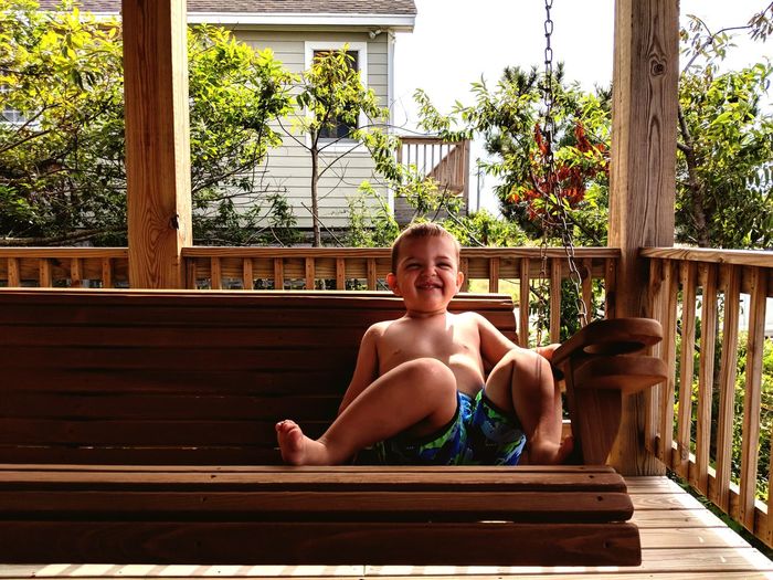 Portrait of shirtless boy smiling while sitting on swing at porch