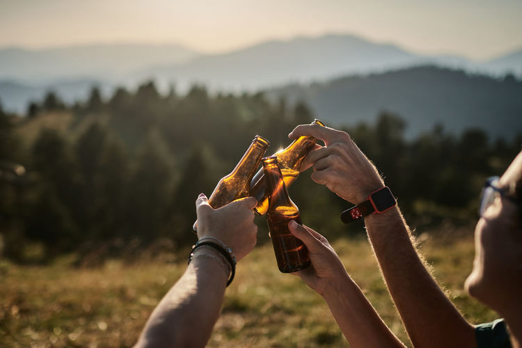 Company of unrecognizable friends relaxing in highland area and clinking bottles of beer while enjoying sunset in summer during weekend