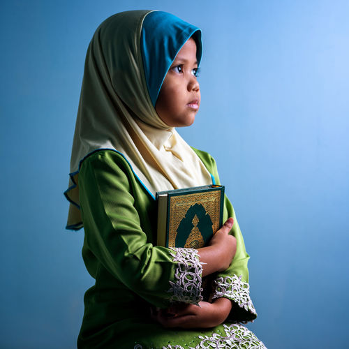 Girl wearing hijab holding koran while sitting against blue background