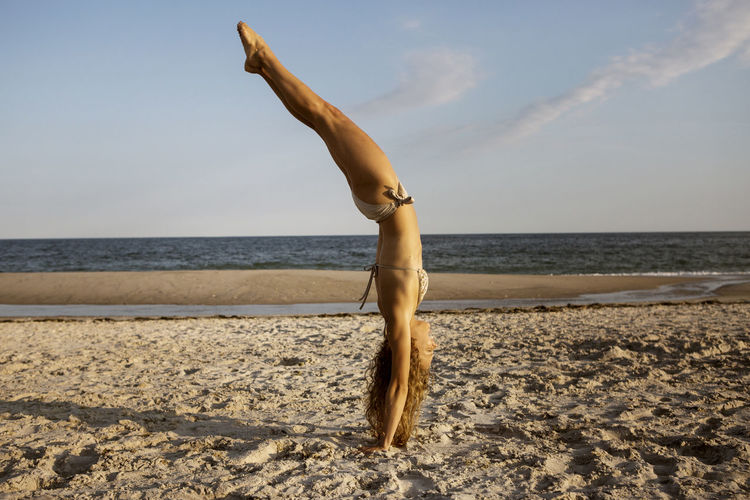 Side view of woman doing handstand at beach against sky