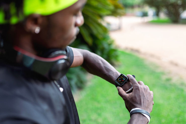 Stylish young african american male runner with headphones on neck checking sport results on fitness tracker during training in green tropical park