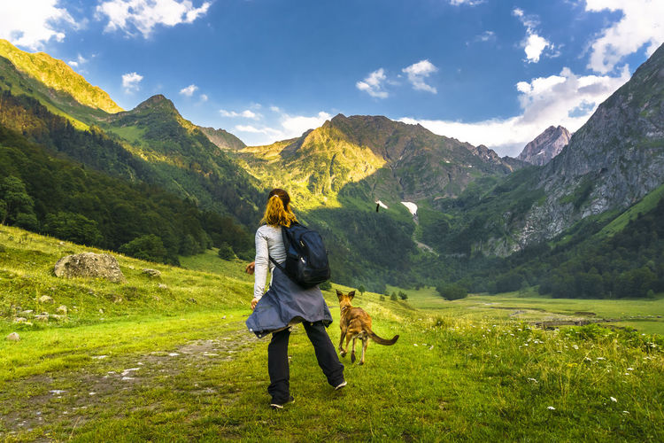 Rear view of woman standing on grass with dog running against mountains and sky