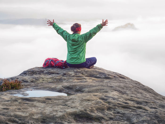 Hiker with backpack sitting on top of a mountain with raised hands and enjoying sunrise