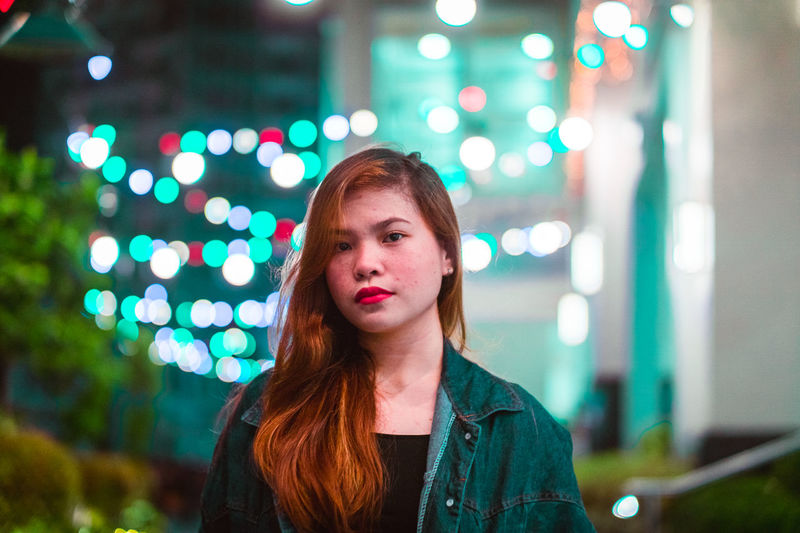 Portrait of young woman with illuminated string lights in background at night