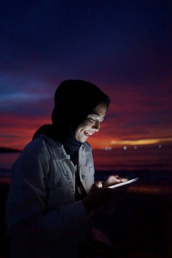 Side view of woman using smart phone against sky at sunset