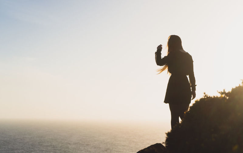 Silhouette woman standing on mountain by sea against clear sky