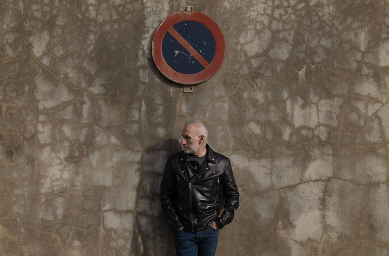 Adult man in leather jacket standing against wall with no parking road sign