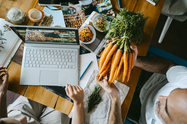 Male colleague holding bunch of carrots by entrepreneur with laptop on table