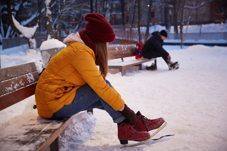 Young woman in a yellow jacket puts on or takes off ice skates while sitting on a bench 