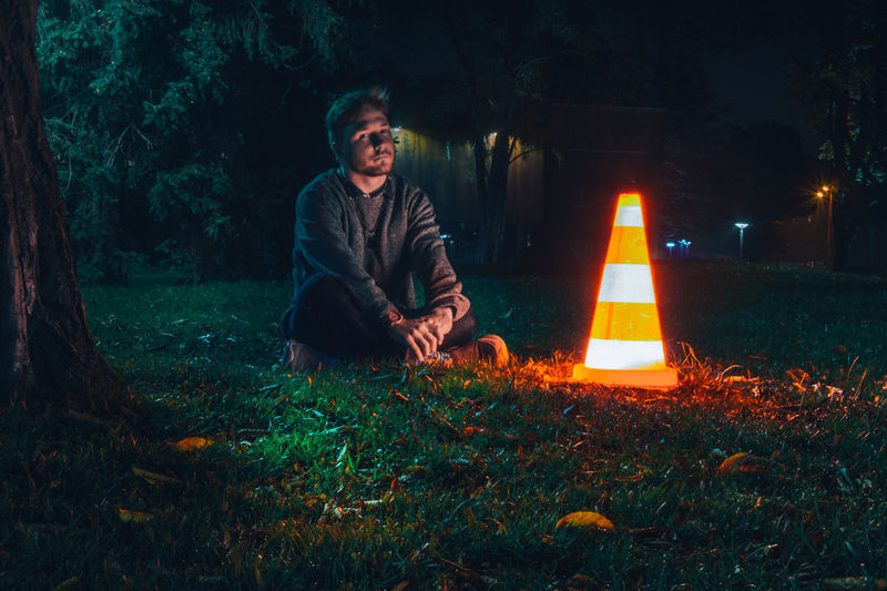 Man sitting on grass by illuminated traffic cone at night