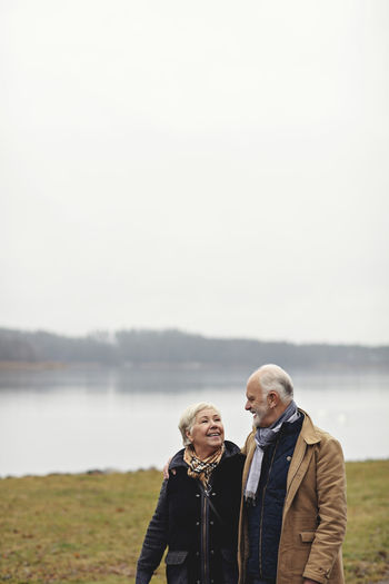 Smiling senior couple looking at each other while standing by lake against clear sky