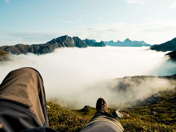 Low section of man sitting on mountain against cloudscape