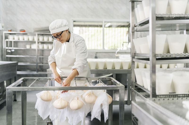 Skilled female chef tying cheese wrapped in clothes on rod at storage room