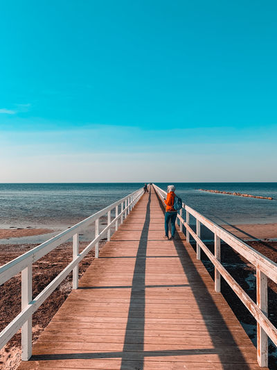 Man standing on pier leading towards sea against blue sky