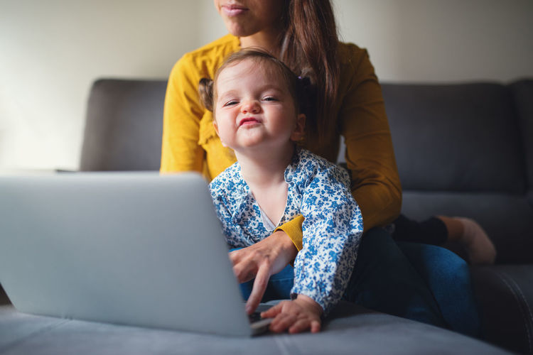Crop unrecognizable busy mother hugging cute toddler daughter while sitting on sofa and doing remote project on laptop at home