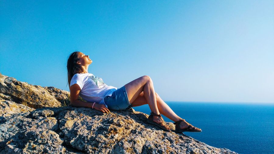 Woman lying on rock by sea against clear blue sky