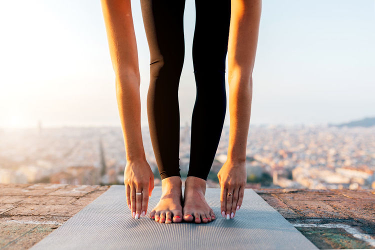 Crop unrecognizable barefoot female in leggings performing uttanasana pose while practicing yoga on mat on rooftop