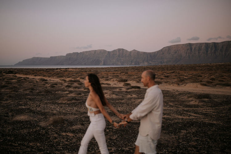 Side view of blurred peaceful couple holding hands while walking in white outfit together in savanna