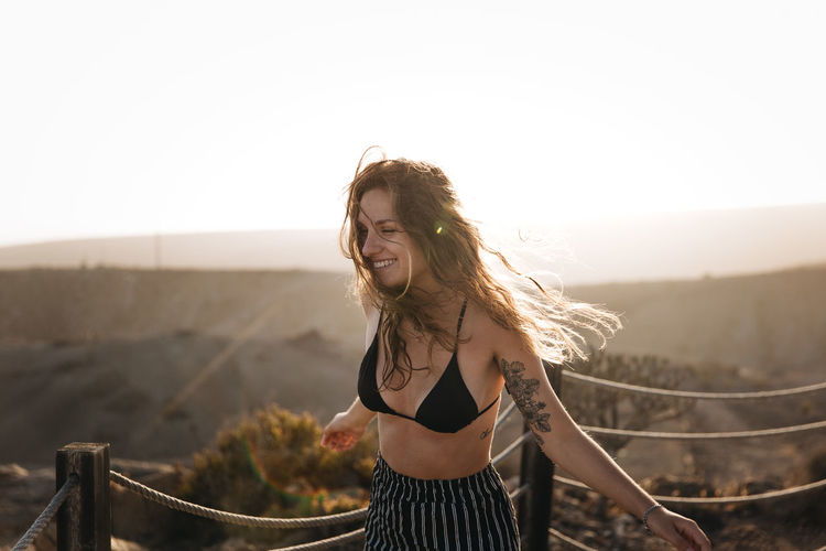 Smiling young woman wearing bikini top against clear sky during sunset