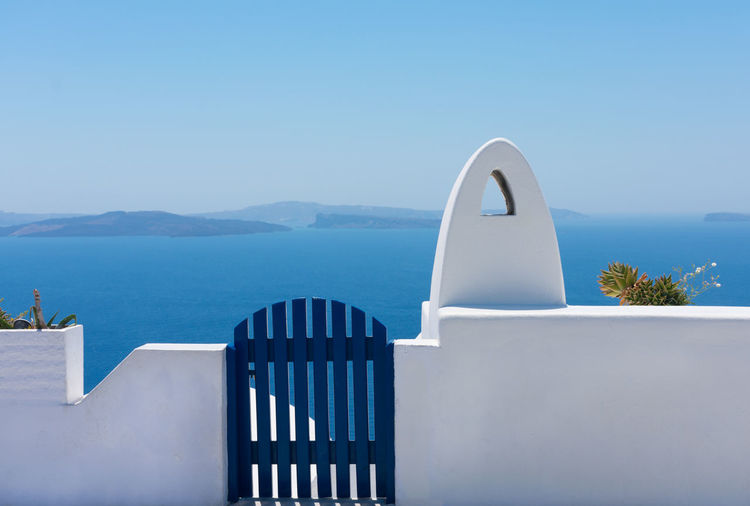 Facade of brick fence or small white wall with a blue gate to enter or exit and a white chimney without smoke with the blue aegean sea at the back in the idyllic island of santorini in greece