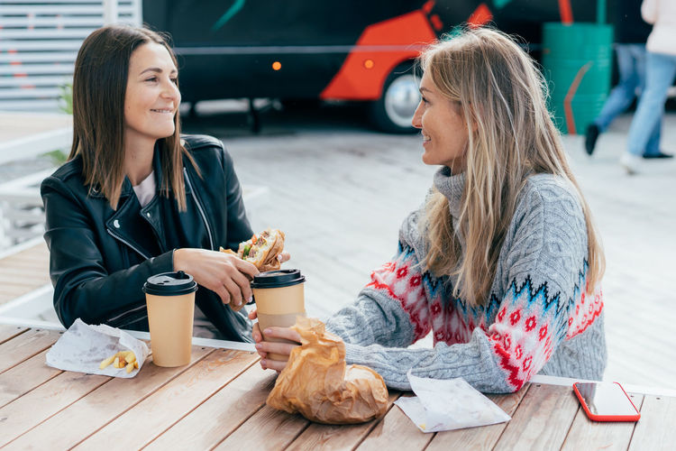 Two cheerful women eat fast food, drink coffee and chat on the street, sitting at the table.