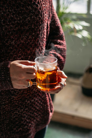 Crop unrecognizable person in warm sweater with glass cup of steaming tasty hot tea standing in light room at home person