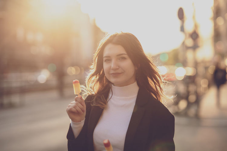 Portrait of young woman holding flavored ice while standing on road in city during sunset