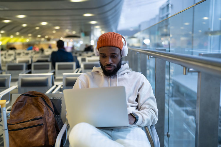 Young hipster black man sitting in airport terminal using laptop wear headphones and listening music