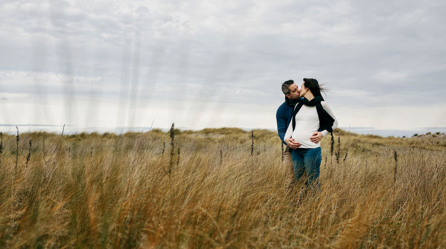 Couple kissing while standing on grassy field against cloudy sky