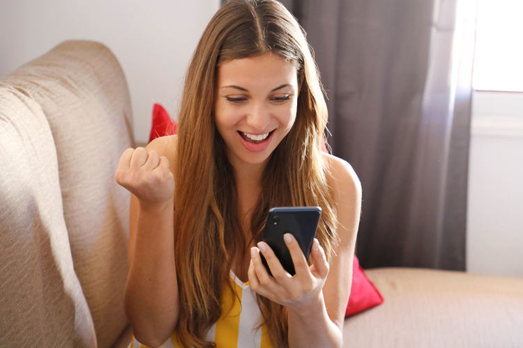 Smiling woman holding smart phone sitting on sofa at home