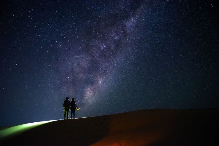 SILHOUETTE PEOPLE STANDING ON FIELD AGAINST STAR TRAILS