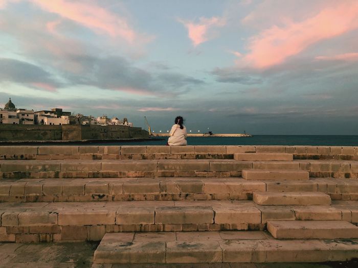 Rear view of girl sitting on staircase by sea against cloudy sky