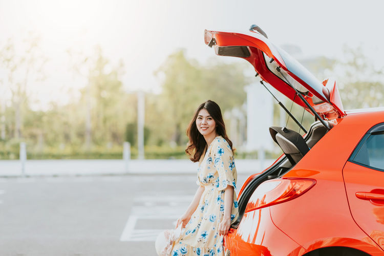 Portrait of smiling beautiful woman sitting on red car trunk