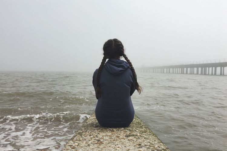 Rear view of teenage girl sitting on pier over sea against clear sky