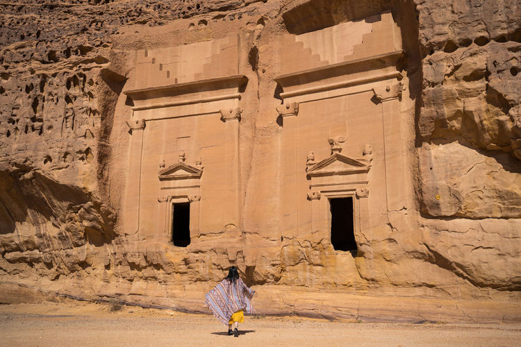 Back view of unrecognizable woman traveler standing near tombs carved into cliffs in madain saleh in saudi arabia