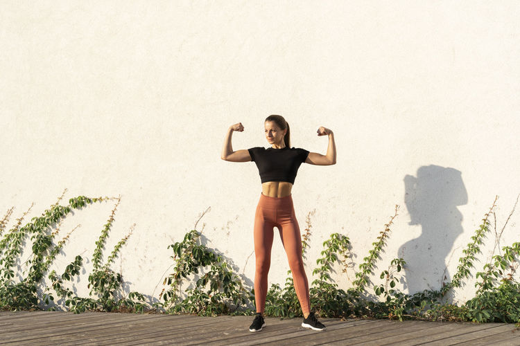 Young female athlete flexing muscle while standing against wall during sunrise