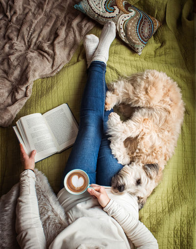 Overhead view of woman's torso on a bed with a book, coffee and a dog.