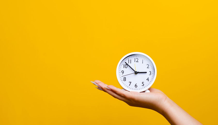 Cropped hand holding clock against yellow background
