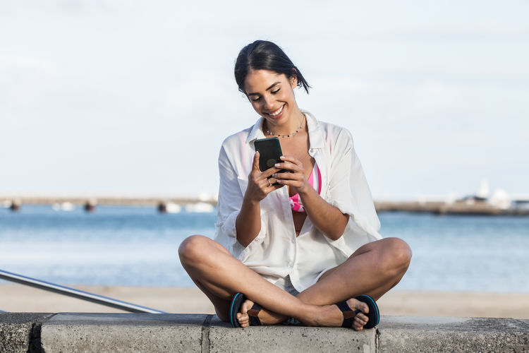 Smiling woman using mobile phone while sitting against bay