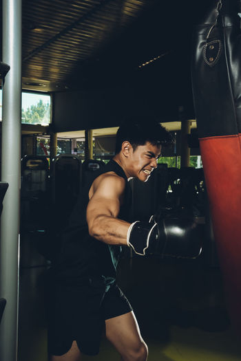 Young focused asian man training boxing performing punches while exercising with heavy punching bag in a modern gym