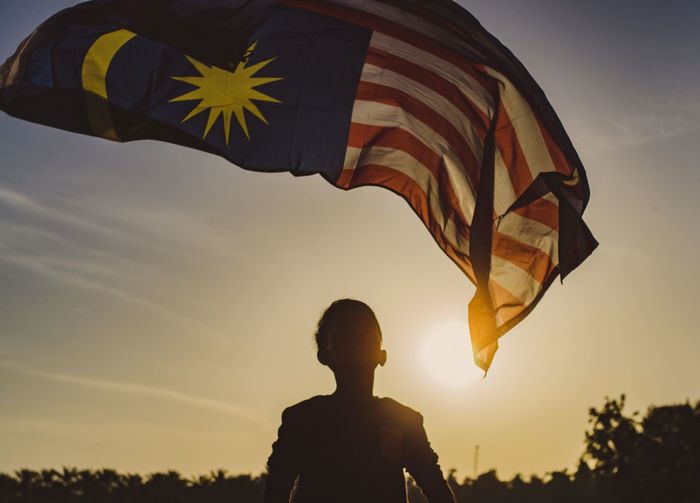 Rear view of boy holding malaysian flag against sky during sunset