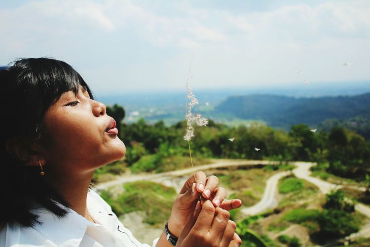 Side view of young woman blowing dandelions against landscape
