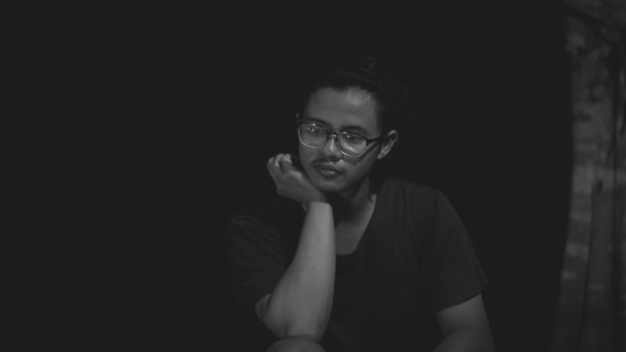 Thoughtful young man wearing eyeglasses sitting against black background