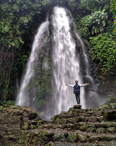Young man with arms outstretched standing on rock against waterfall
