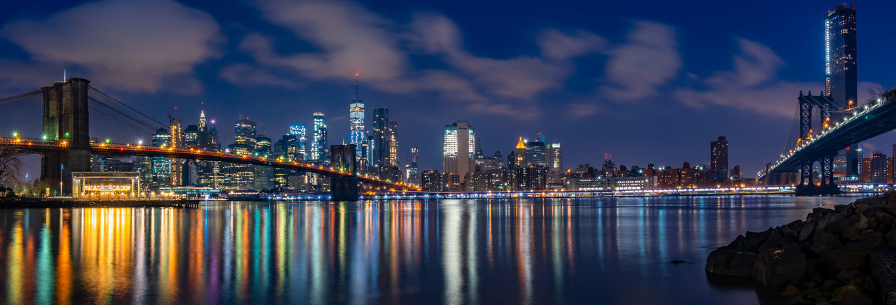Illuminated bridge and buildings by river against sky at night
