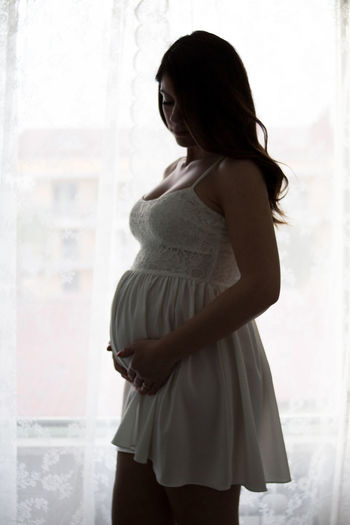 Side view of a young pregnant woman standing against window 