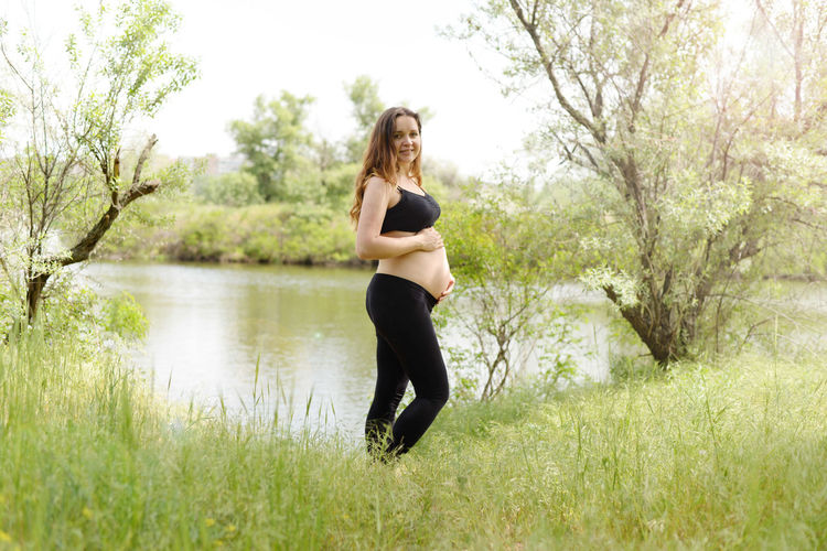 Portrait of pregnant young woman touching abdomen while standing on grass