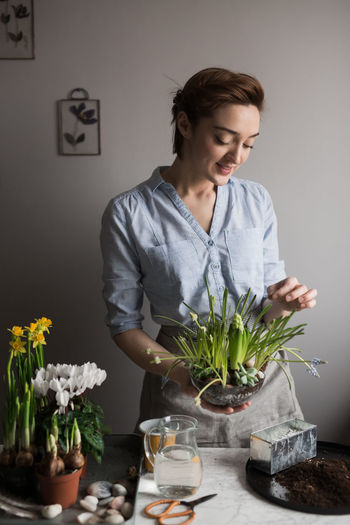 Content female gardener in apron standing with assorted potted fresh flowers and plants at table at home and looking down