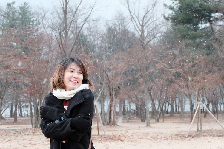 Cheerful woman hugging self while standing against trees at park during snowfall