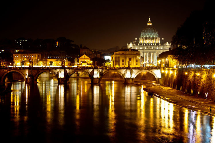 Illuminated st peter basilica by bridge and river against sky at dusk